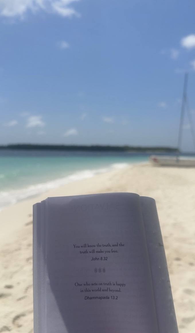 An open book propped on the sand, sea and a sailboat behind