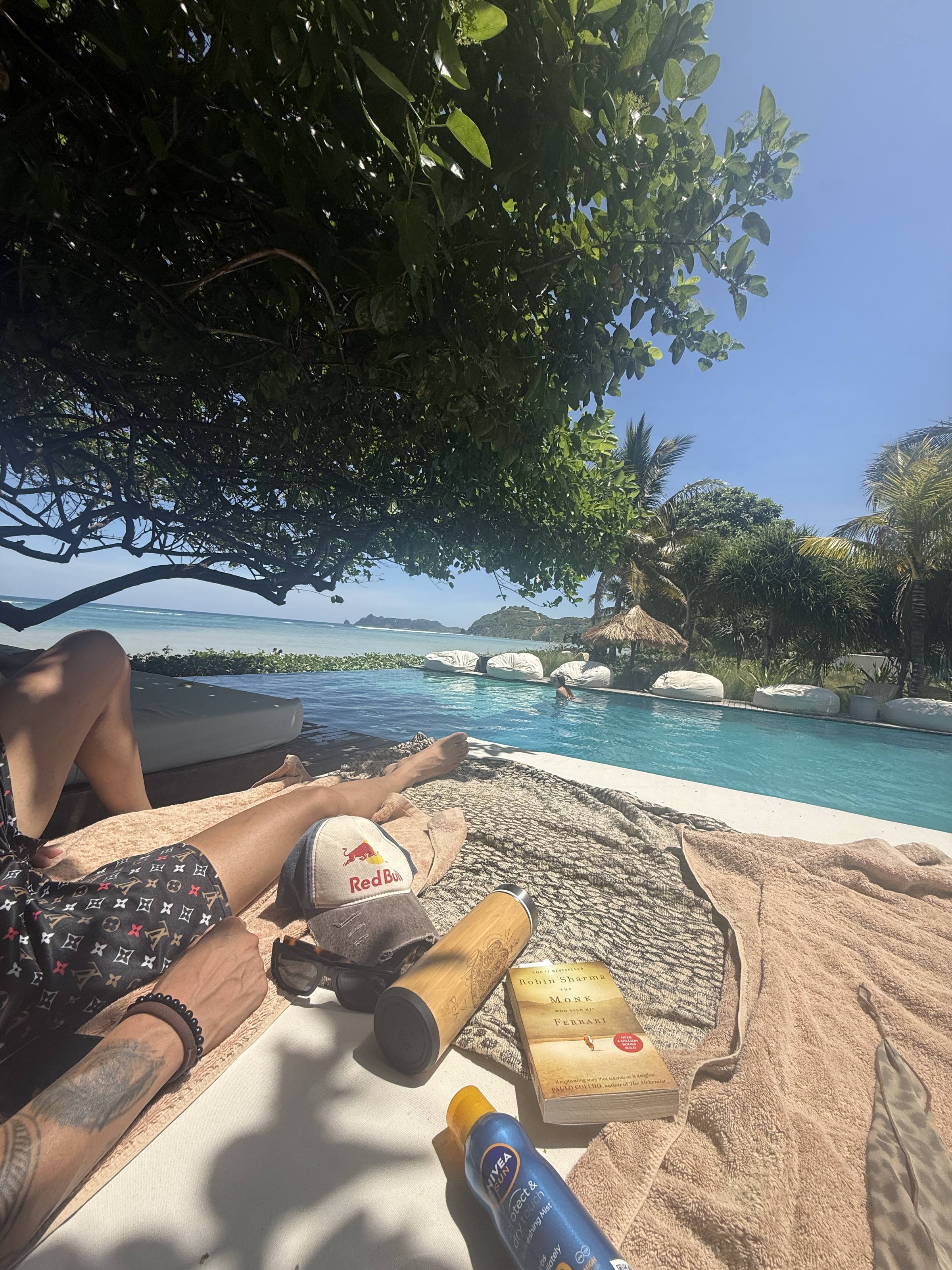 A paperback on a towel beside a flask and sunglasses, pool and ocean behind
