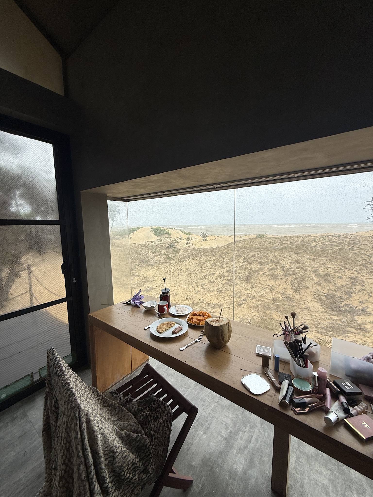 Breakfast and a makeup bag spread on a desk facing dunes and the sea